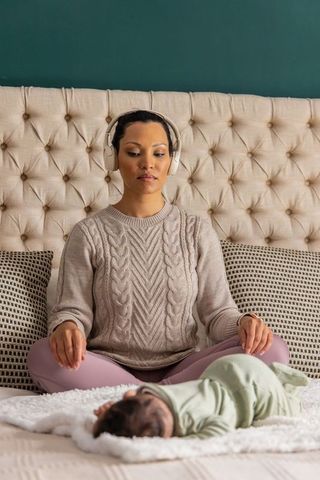 Parent meditating with headphones beside sleeping baby on bed, cozy bedroom mindfulness