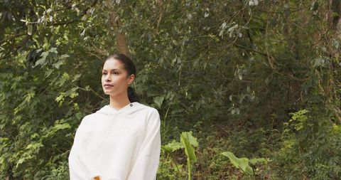 Young woman standing in lush forest wearing white hoodie and gold hoop earrings, serene