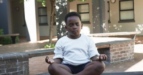 Young black boy meditating on brick ledge in sunlit courtyard
