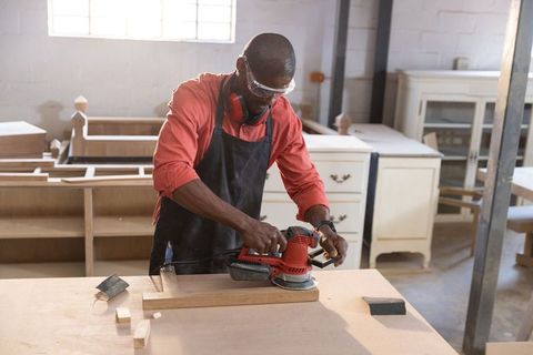 Carpenter sanding hardwood plank with red orbital sander in sunlit woodworking shop