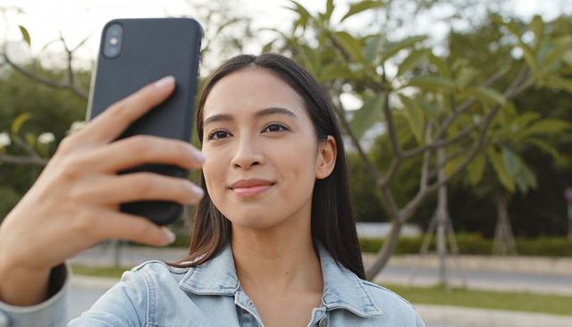 Asian woman taking selfie on tree-lined park path wearing denim jacket, natural portrait