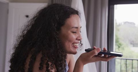 Smiling african american woman sending voice message on smartphone at sunny balcony window