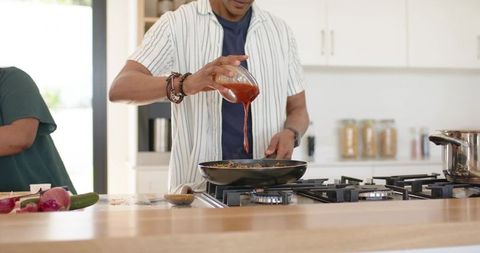 Couple cooking on modern kitchen island pouring tomato sauce into sizzling frying pan