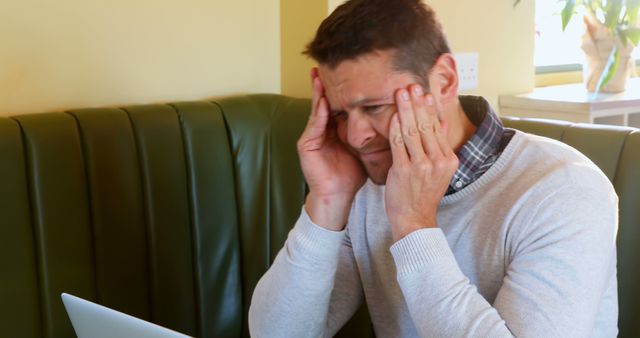 A man holds his head in frustration while using a laptop in a stylish cafe. This can be used in stories or advertisements about work-related stress, technical issues, or challenges faced while working remotely.