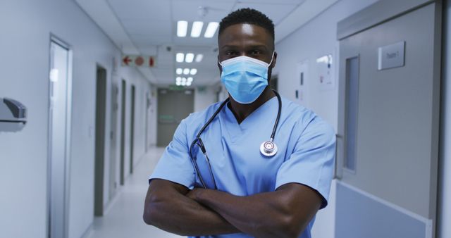 Portrait of african american male doctor wearing face mask and scrubs standing in hospital corridor. medicine, health and healthcare services during coronavirus covid 19 pandemic.