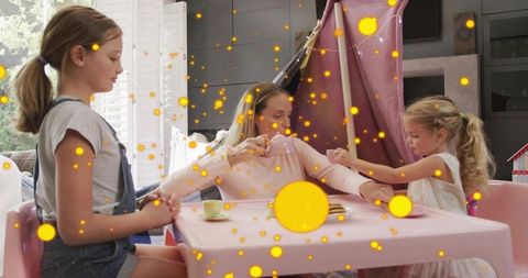 Mother pouring tea for daughters at pink play table inside cozy living room blanket fort
