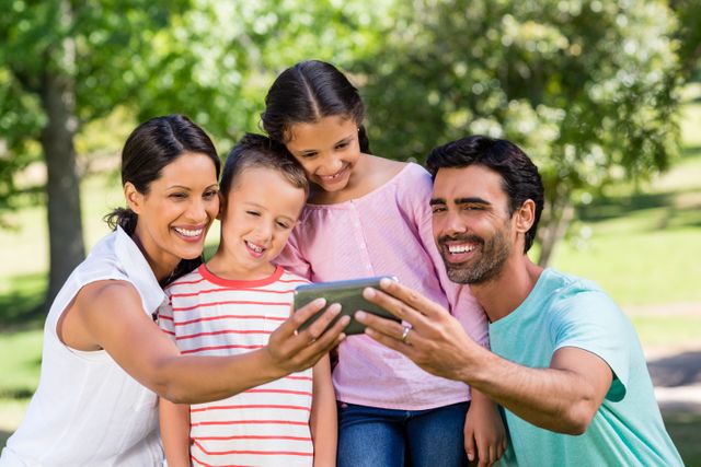 Family enjoying a sunny day in the park, capturing a moment with a mobile phone. Perfect for themes related to family bonding, outdoor activities, summer fun, and technology use in everyday life.