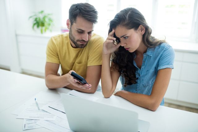 Young couple sitting at kitchen table, looking concerned while reviewing bills and using laptop. Ideal for illustrating financial stress, budgeting, home finances, and relationship challenges. Suitable for articles, blogs, and advertisements related to personal finance, financial planning, and family life.