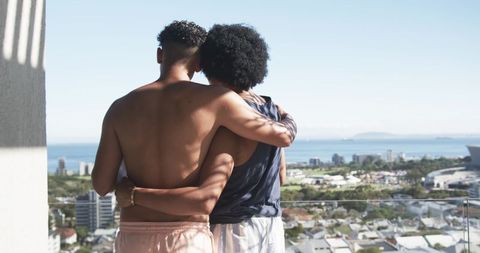 Couple embracing on sunny balcony overlooking ocean and city skyline, rooftop sunlight