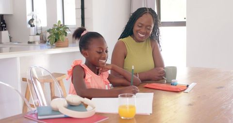 African american mother smiling while helping daughter with homework at sunlit kitchen table