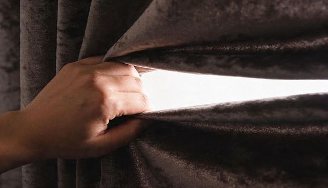 Adult hand pulling velvet curtain revealing narrow backlit light slit and textured drapery