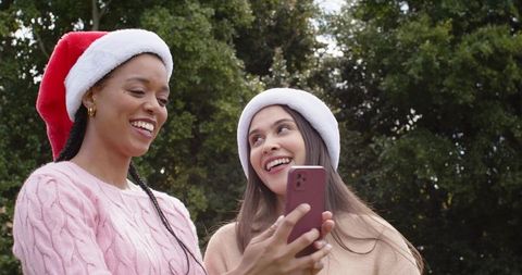 Friends wearing santa hats sharing smartphone and smiling in park holiday lifestyle