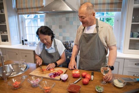 Senior asian couple cooking together in modern kitchen chopping tomatoes and sharing wine