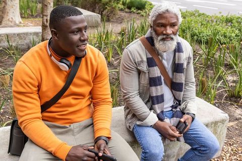 African american friends sitting on urban bench chatting using smartphones at city plaza