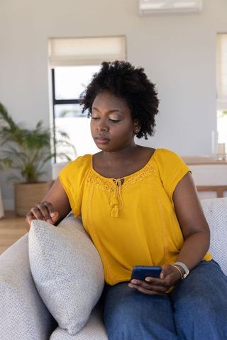 African american woman relaxing on sofa using smartphone in bright home living room