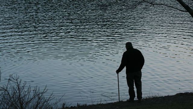 Senior man standing at lakeside steadying with cane at dusk, silhouette mood of solitude