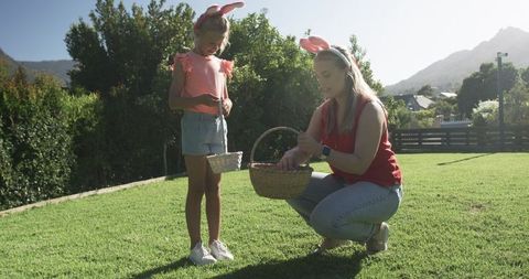 Mother and daughter wearing bunny ears collecting eggs on sunlit backyard lawn