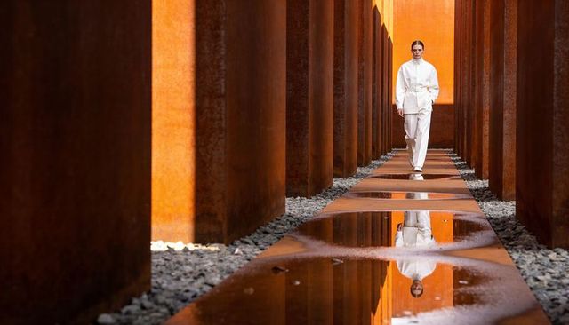 Model walking through corten steel corridor with mirror puddles and minimalist outfit