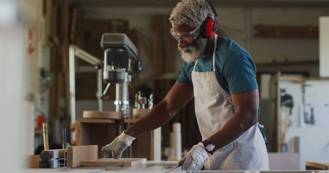 Senior male carpenter wearing safety gear including goggles, ear protection, and gloves working in a woodworking workshop. He is focused on cutting wood pieces, demonstrating craftsmanship and attention to detail. Useful for concepts of skilled labor, woodworking hobbies, safety practices, and portraying an experienced artisan at work.