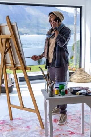 Artist painting on easel while talking on phone in bright waterfront studio