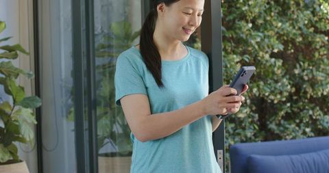 Smiling asian woman checking smartphone in doorway on sunny patio with plants and couch