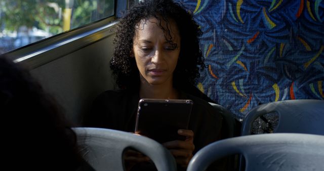 This image depicts an African American woman with curly hair using a tablet while riding a bus. The concept highlights the use of technology in daily commutes via public transportation. This can be useful for articles or advertisements focused on public transit systems, technology integration in everyday life, digital literacy, or urban commuting. Ideal for blogs, promotional materials, and editorial use.
