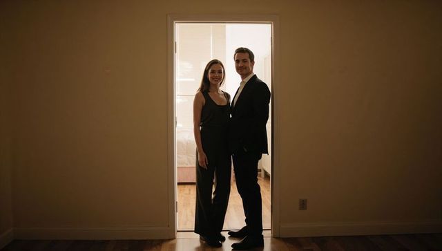 Couple standing in narrow doorway wearing formal attire backlit by bedroom window light