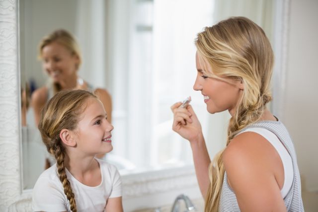Mother and daughter sharing a joyful moment while applying lipstick in the bathroom. Perfect for themes related to family bonding, parenting, beauty routines, and mother-daughter relationships. Ideal for use in advertisements, blogs, and articles focusing on family life, beauty tips, and parenting advice.