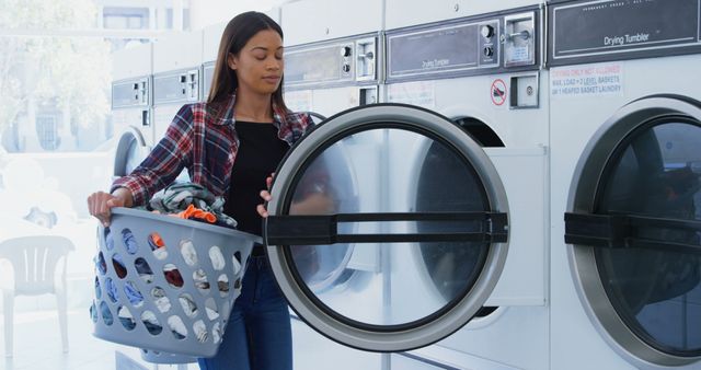 This image shows a woman dressed casually, holding a laundry basket and loading a washing machine at a laundromat. Great for illustrating concepts related to daily routines, cleanliness, domestic chores, laundry services, or community facilities. It can be used in articles or advertisements focused on lifestyle, laundry products, or public services.