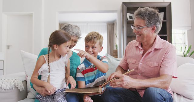 Happy caucasian grandparents and grandchildren sitting on sofa and looking at photo album pictures. Lifestyle, domestic life, family, and togetherness.