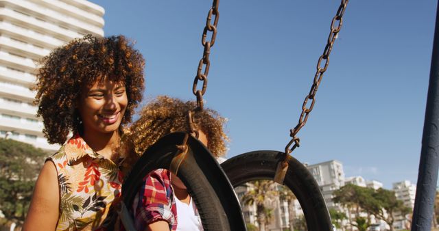 Happy biracial mother pushing son playing on swing at sunny playground, copy space. Motherhood, childhood, togetherness, summer, vacations, fun and free time, unaltered.