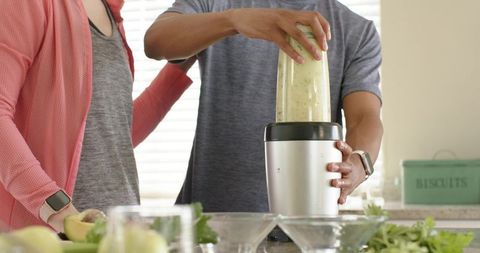 Couple preparing green smoothie with personal blender on sunlit kitchen countertop
