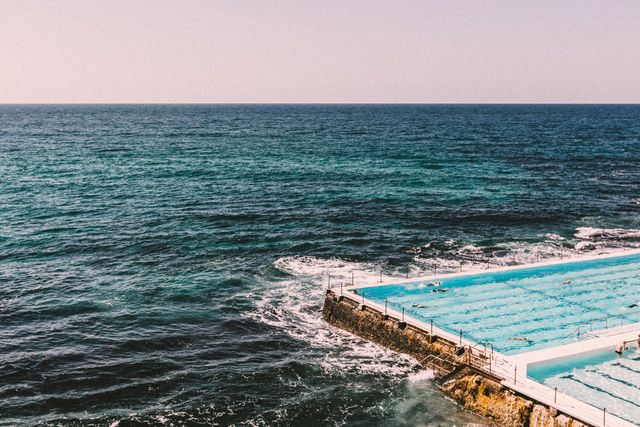Ocean swimming pool placed next to open sea with gentle waves under clear sky. Suitable for travel ads, vacation blogs, leisure activity websites, and outdoor lifestyle promotions.