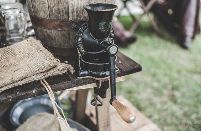 Shows an old-fashioned, manual coffee grinder on a rustic wooden table outdoors, with a burlap sack and other traditional elements. Ideal for themes related to heritage, nostalgia, traditional coffee-making methods, vintage kitchen tools, outdoor events, and antique equipment enthusiasts.