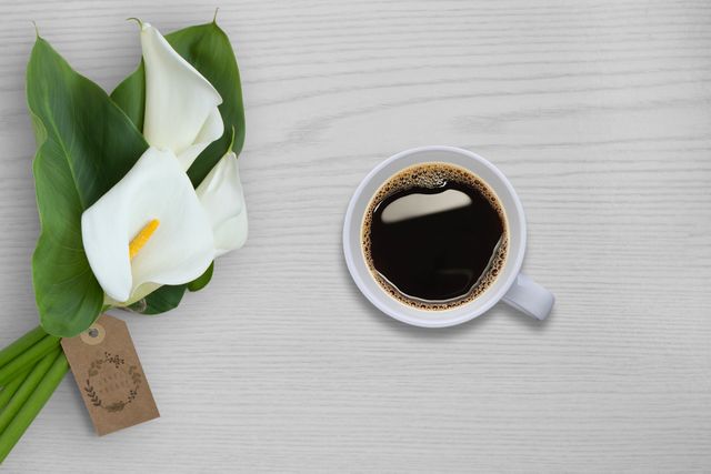 Cup of black coffee next to a bouquet of white calla lilies on a light wooden table. Perfect for themes of morning routines, relaxation, and freshness. Ideal for blogs, social media content, and print advertising focusing on coffee, florals, and minimalistic lifestyle aesthetics.