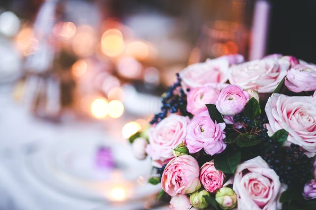 Romantic wedding centerpiece featuring pink roses and ranunculus with warm bokeh lights in background, perfect for wedding invitations, decor blogs, florists' promotional materials, event planning guides, and romantic-themed projects.