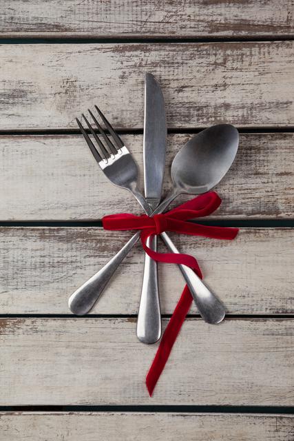 This image features a set of cutlery, including a fork, knife, and spoon, tied together with a red ribbon on a rustic wooden plank background. Ideal for use in dining-related content, kitchen decor ideas, festive table settings, and celebration themes.