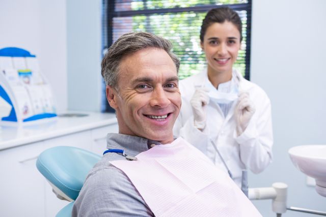 Man sitting in dental chair smiling with dentist standing in background. Suitable for use in healthcare, dental care, and medical treatment contexts. Ideal for illustrating dental checkups, oral health, and professional dental services.