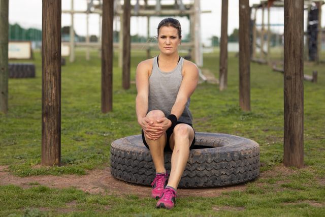 Portrait of a Caucasian woman wearing sports clothes at an outdoor gym sitting on a tyre, holding her knee with both hands and stretching before a bootcamp training session