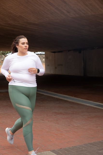 Curvy woman with long dark hair running in an urban pedestrian area, wearing sports clothes and earphones. Ideal for promoting fitness, active lifestyle, sportswear brands, health and wellness campaigns, and urban exercise routines.