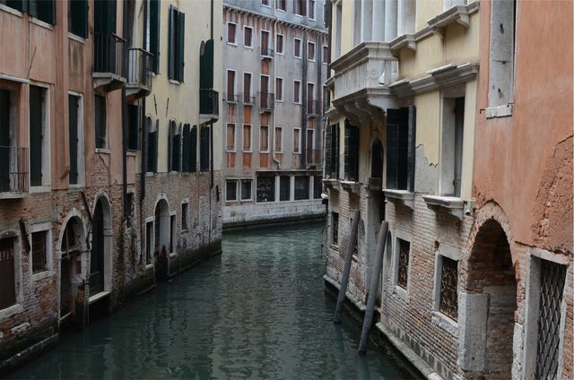 Narrow Venice canal between weathered buildings with bricked facades and arched doorways. Perfect for travel guides, tourism advertisements, architectural studies, and content on European travel destinations.