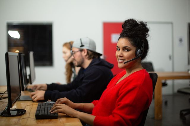 Side view of a biracial woman working in a creative office, sitting by a desk, with a computer on it, wearing a wireless headset, writing on a keyboard, looking at the camera, with her coworkers in the background