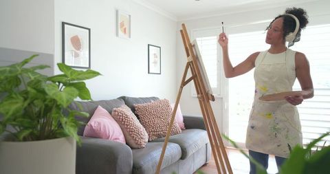 African american woman painting at home studio wearing apron headphones holding palette