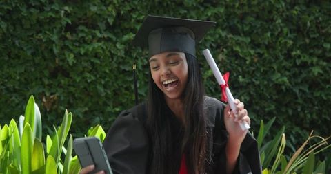 Young asian woman celebrating virtual graduation outdoors cap and gown holding diploma