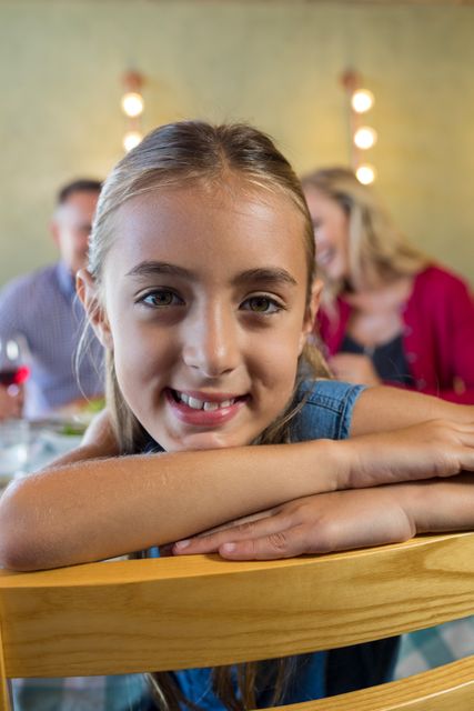 Smiling girl leaning on chair with family in background at restaurant. Ideal for use in family-oriented advertisements, restaurant promotions, or articles about family dining experiences. Highlights themes of togetherness, joy, and casual dining.