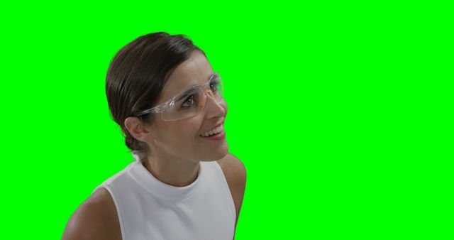 Young woman wearing safety glasses and white top, smiling against green background. Ideal for use in advertisements for safety equipment, industrial training material, or promotional content showcasing optimism and professionalism in work safety practices. Suitable for both digital and print media.