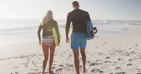 Active couple walking with surfboards on beach shoreline