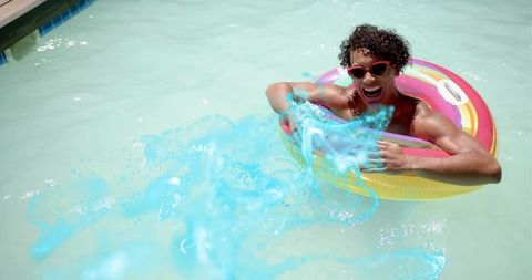 Young man floating on colorful inflatable ring in sunny pool, smiling and splashing