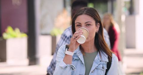 Young woman sipping takeaway coffee while walking urban sidewalk with backpack