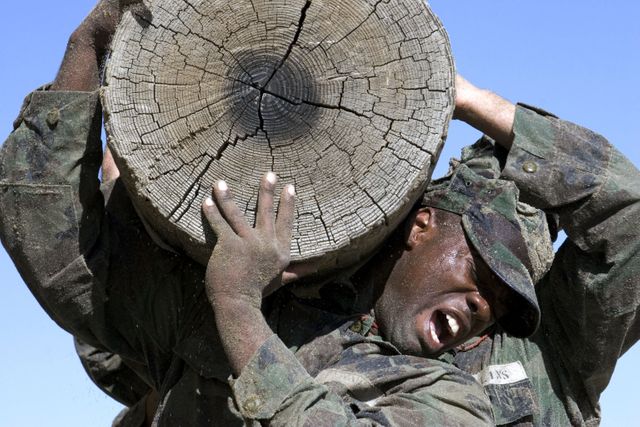 Soldier in camouflage lifting heavy wooden log during intense military training exercise, showcasing physical endurance and teamwork. Useful for illustrating concepts of strength, determination, military training, teamwork, and physical challenges. Ideal for use in articles, blogs, advertisements related to military, fitness, and team-building events.